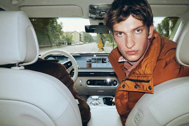 Young man in an orange puffer jacket looking back over the front seat inside a modern car interior.