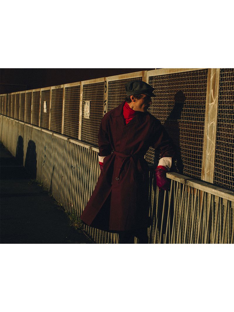 Model Clement Chabernaud wearing a burgundy belted trench coat and a flat cap while leaning against a metal mesh fence in sunlight.