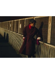 Model Clement Chabernaud wearing a burgundy belted trench coat and a flat cap while leaning against a metal mesh fence in sunlight.