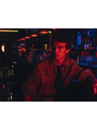 Model Clement Chabernaud sits at a dimly lit bar wearing a plaid jacket and necktie under vibrant red lighting.