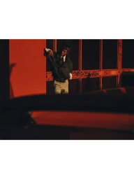 Model Clement Chabernaud leans against a vibrant red exterior gate in warm golden light wearing a dark jacket and light trousers.