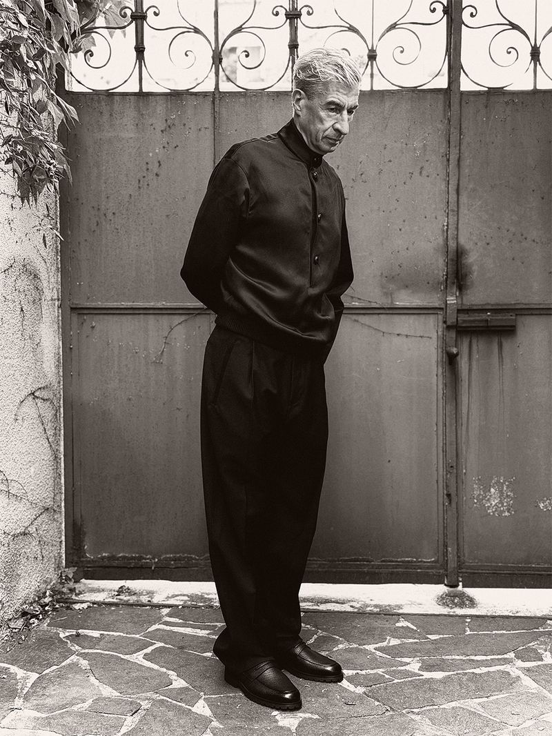 Maurizio Cattelan in a black buttoned jacket and pleated trousers standing before a metal gate with hands behind his back.