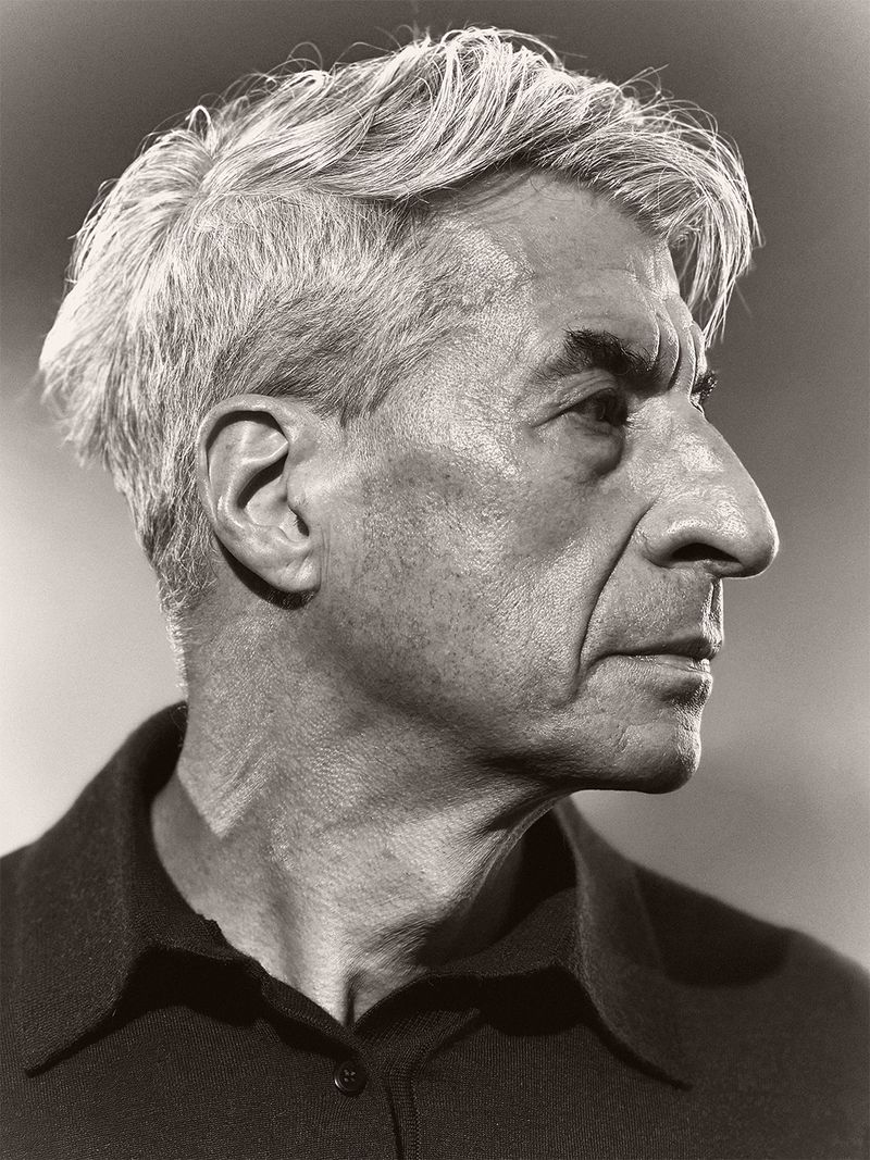 Profile portrait of artist Maurizio Cattelan in a dark collared shirt against a neutral studio backdrop.