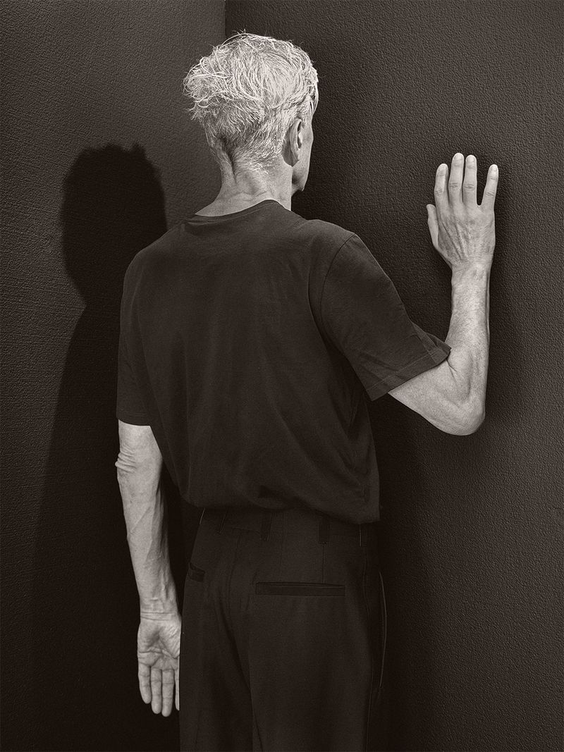 Man with short grey hair seen from behind facing a dark corner with his right hand flat against the textured wall.