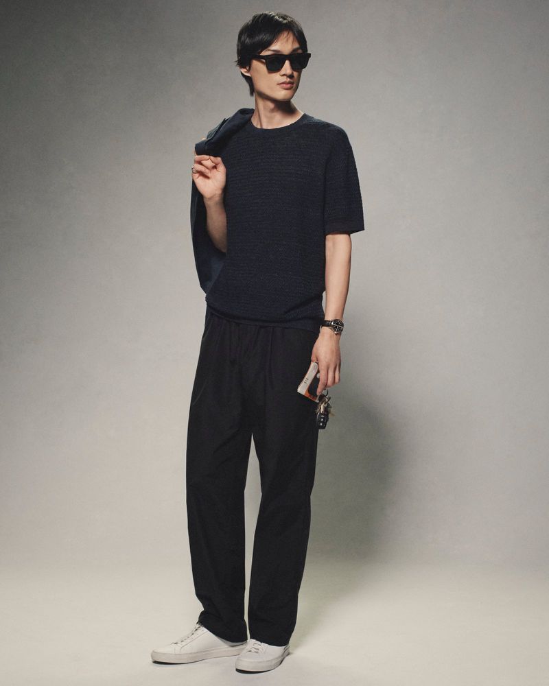 A stylish Asian man in a dark knit shirt and black pants stands in a studio setting, with a jacket slung over his shoulder.