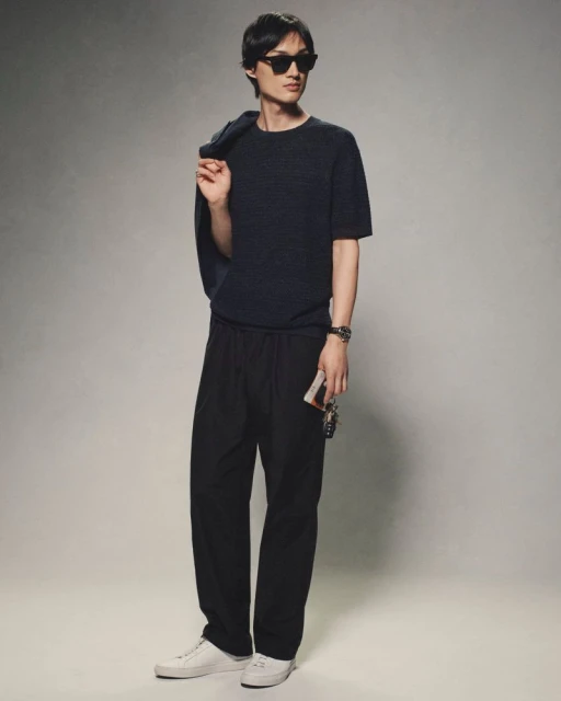 A stylish Asian man in a dark knit shirt and black pants stands in a studio setting, with a jacket slung over his shoulder. 