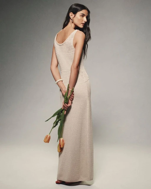 Elegant woman in a long knitted dress holding a bouquet of tulips, posing in a photo studio with a soft, neutral backdrop.