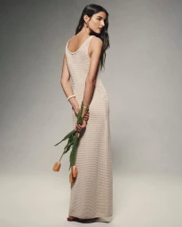 Model looking over her shoulder in a cream sleeveless knit maxi dress holding peach tulips behind her back in a studio setting.