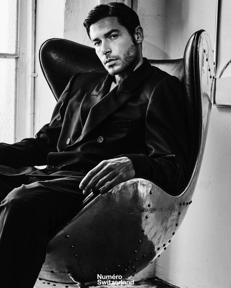 Model Andrea Denver in a dark double-breasted suit seated in a metallic egg chair for Numéro Switzerland.