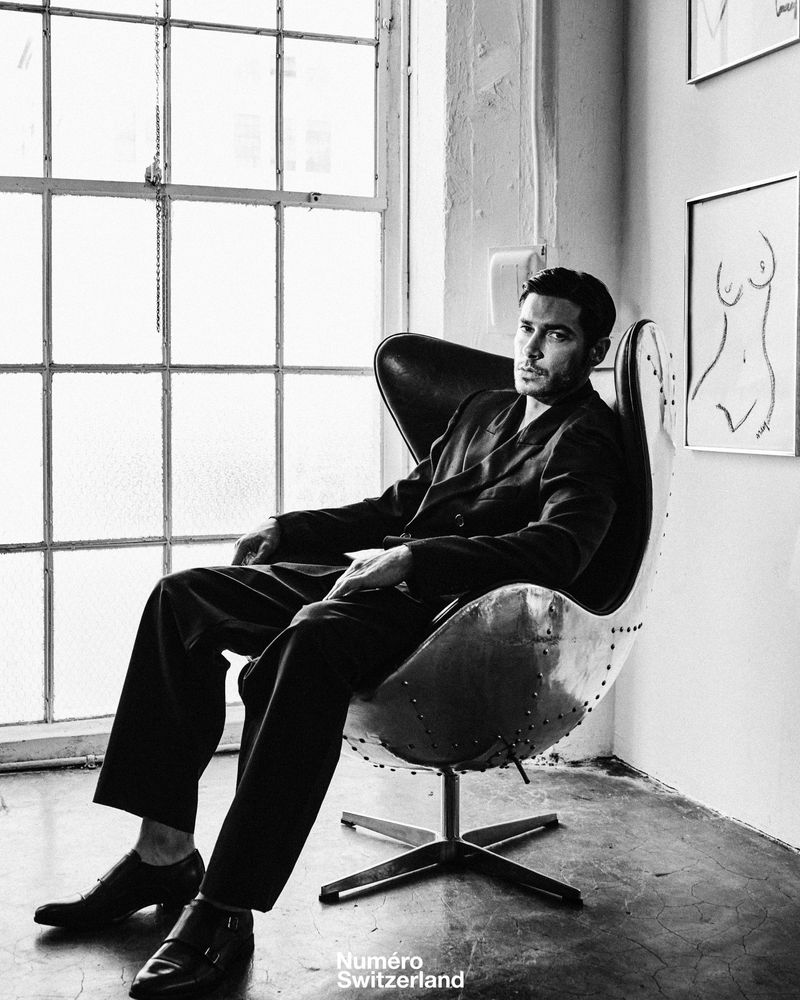 Andrea Denver wearing a dark suit seated in an aviator egg chair beside a multi-pane window in black and white.