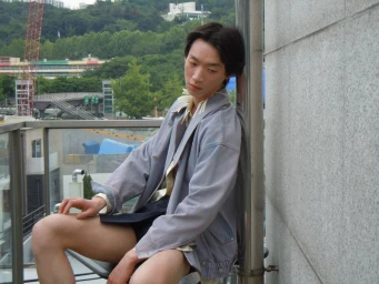 Model Seungmin We seated on a balcony wearing an oversized light blue denim jacket with an urban hillside background.