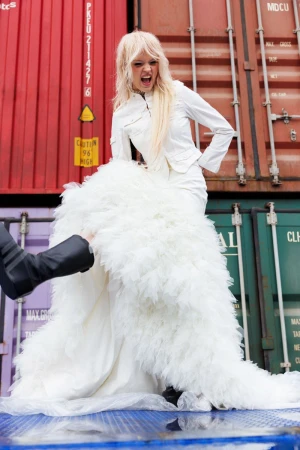 Model Batura Aliaksandra shouting while wearing a dramatic white ruffled gown and jacket in front of shipping containers.