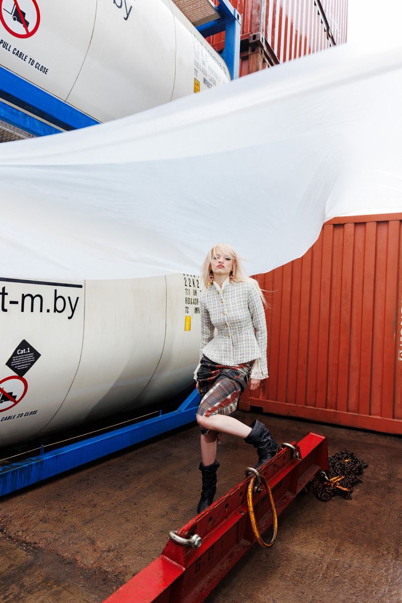 Model posing in a light tweed jacket and plaid skirt atop a red steel beam in an industrial shipping container yard.