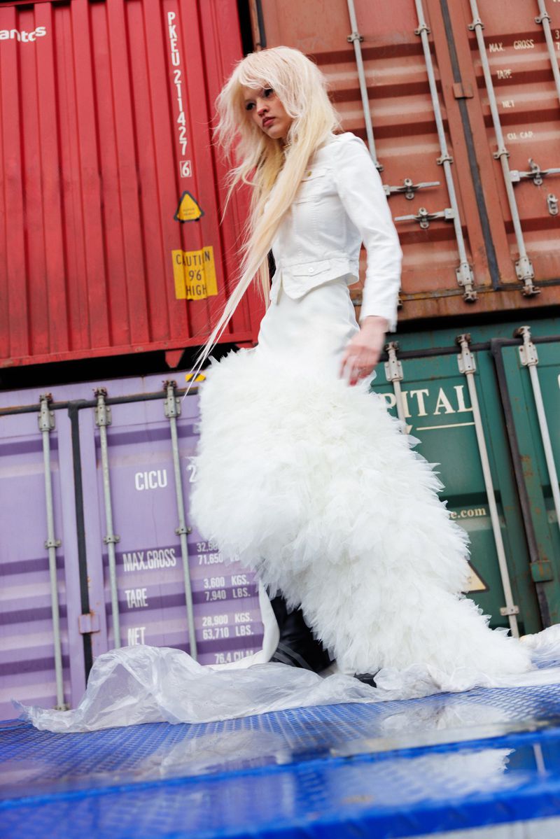 Model Batura Aliaksandra wearing a white jacket and feathered skirt standing against stacked industrial shipping containers.