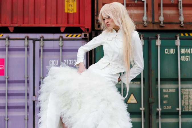 Model with long blonde hair in a white jacket and voluminous layered white skirt posing against stacked shipping containers.
