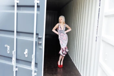Model Batura Aliaksandra wearing a floral sheer dress, pink opera gloves, and red heels inside an industrial shipping container.