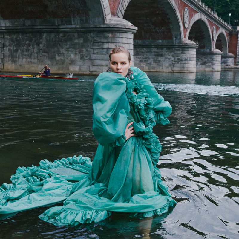 Eva Herzigova poses in river water wearing a voluminous ruffled green gown in front of an arched stone bridge.