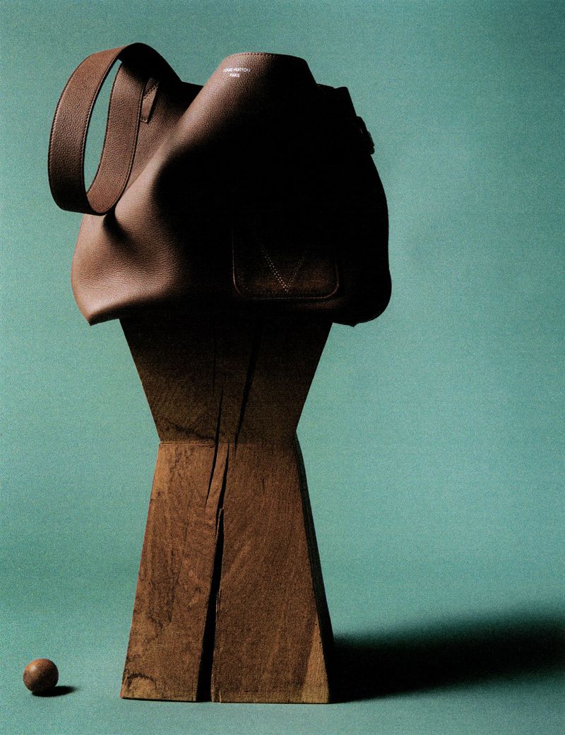 A brown leather handbag rests atop a raw, weathered wooden post against a teal background with a small wooden ball on the floor.