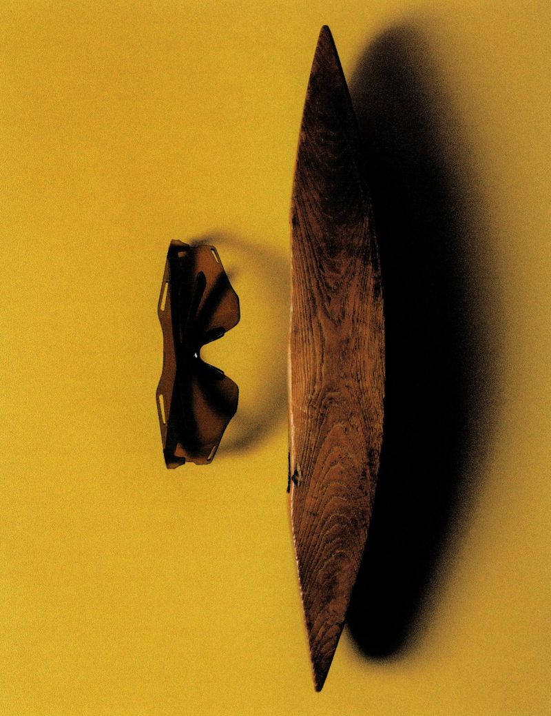 Black shield sunglasses next to an elongated pointed wooden object casting a dark shadow on a yellow surface.