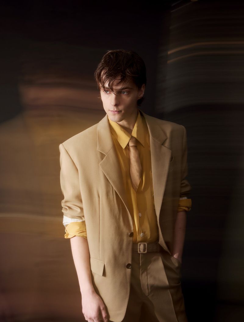 Corey Fogelmanis posing in an oversized beige tailored suit jacket, yellow collared shirt, and matching tie.
