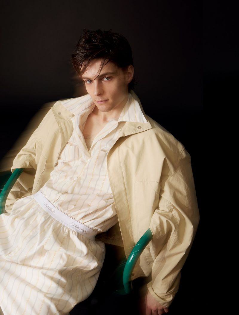 Corey Fogelmanis seated in a green armchair wearing a light beige jacket and striped loungewear against a dark background.