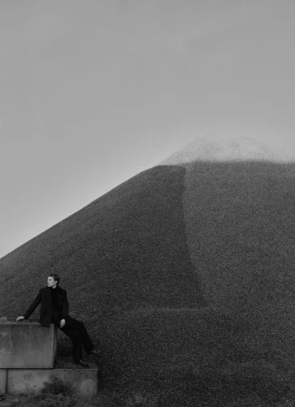 A man in a dark tailored suit seated on stacked concrete blocks beside a massive gravel mound under an overcast sky.