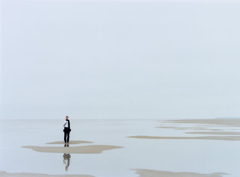 Man standing on a flat sandy tidal beach with reflections in shallow water under an overcast sky.