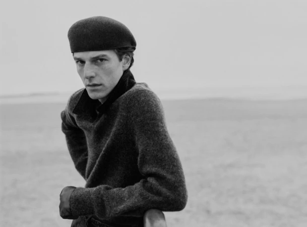 Man in a dark beret and wool knit sweater leaning against a railing in a black and white portrait outdoors.