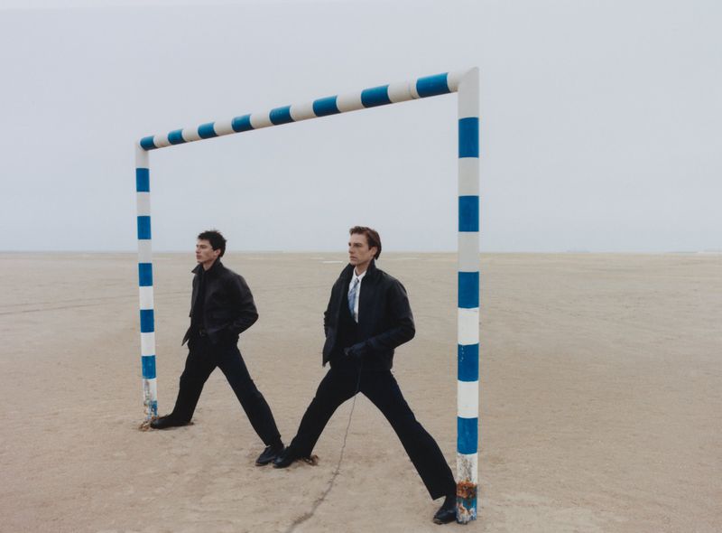 Two male models in dark coats pose under a blue and white striped goal post on a vast sandy plain under an overcast sky.