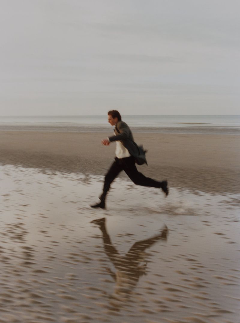Man in a dark coat and trousers running across a wet sandy beach under an overcast sky.