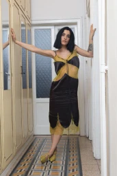 Woman with a dark bob posing in a hallway wearing a black satin and yellow lace midi dress with matching yellow heels.