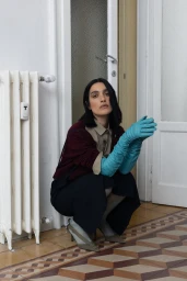Woman with dark hair crouching on patterned tile floor wearing long turquoise leather gloves, a burgundy cardigan, and heels.