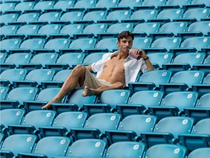 Lorenzo Musetti lounges barefoot across rows of teal stadium seats wearing an unbuttoned white shirt and holding a drink.