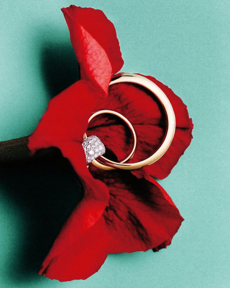 Two intertwined gold rings, one featuring small diamonds, resting on a deep red flower petal against a solid turquoise background.