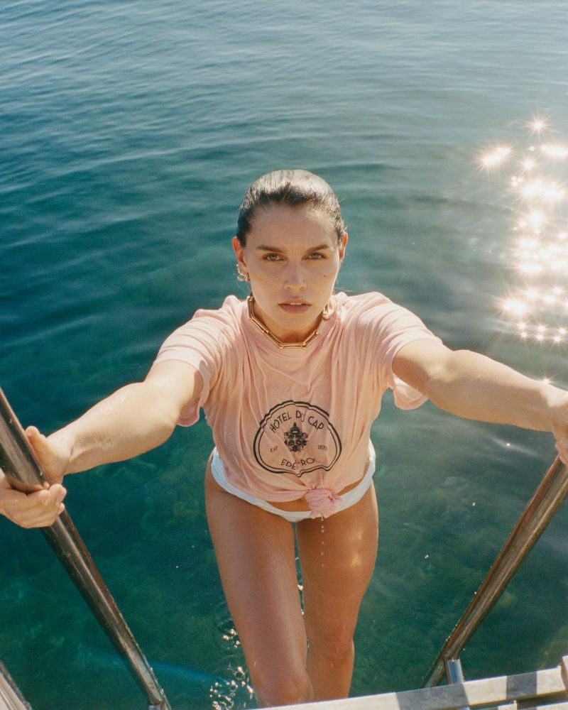 Woman in a pink t-shirt emerges from clear water on stairs, bathed in sunlight, showcasing swimwear fashion and a serene aquatic scene.