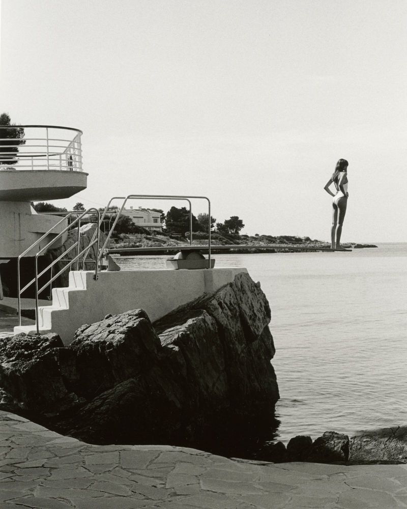 Monochrome image of a woman in a swimsuit standing on a cliff overlooking the sea, with a diving platform visible in the foreground.