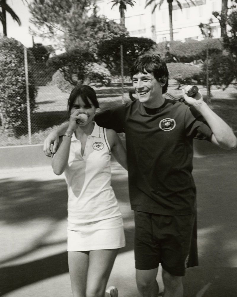 Black and white shot of a smiling couple standing on a tennis court with trees and a building visible in the background on a sunny day.
