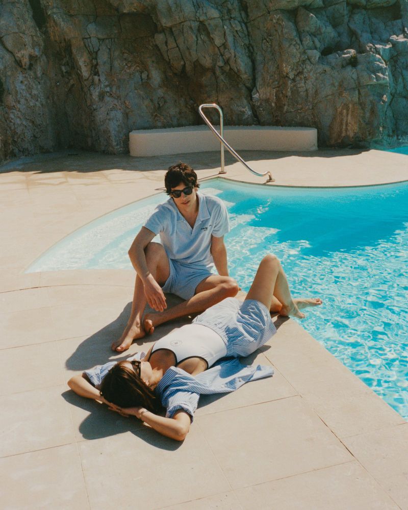 A woman lies on a pool deck while a man sits nearby in matching outfits, showcasing fashion and leisure in an outdoor setting with a rocky backdrop.