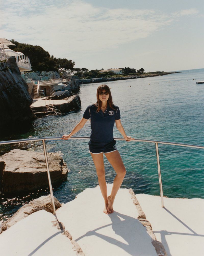 A woman stands on a boat overlooking the ocean, wearing a navy top and shorts, with coastal buildings visible in the background.