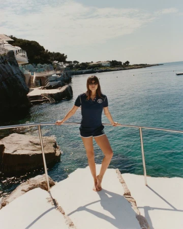A woman stands on a boat overlooking the ocean, wearing a navy top and shorts, with coastal buildings visible in the background.