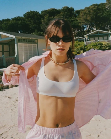 A young woman on a beach in a pink-striped shirt, white top, and pink shorts stands confidently, the blue sky and beach houses behind her.