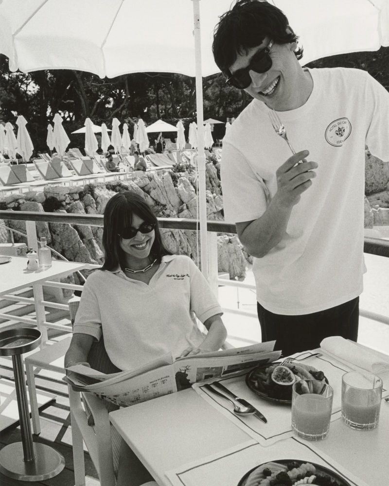 Black and white shot of a fashionable couple seated at an outdoor cafe table with drinks, fruit, and a menu, by a rocky coastline.