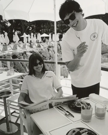 Black and white shot of a fashionable couple seated at an outdoor cafe table with drinks, fruit, and a menu, by a rocky coastline.