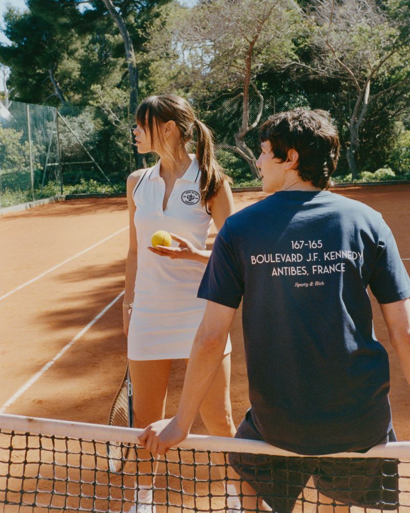 Two people on a tennis court on a sunny day. A woman in a white tennis dress holds a ball, with a man in a blue t-shirt behind her.