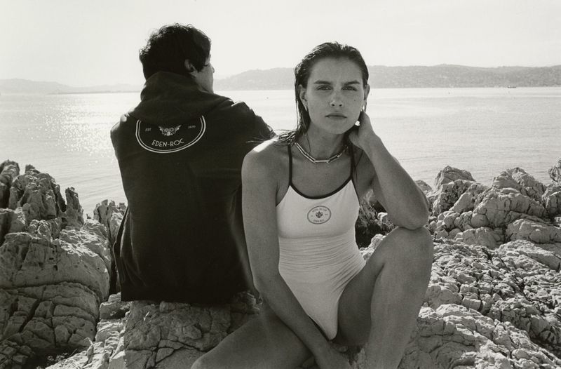 Woman in swimsuit poses on rocky outcrop near man in jacket. Ocean and distant hills in background. Black and white, sunny day.