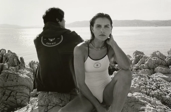 Woman in swimsuit poses on rocky outcrop near man in jacket. Ocean and distant hills in background. Black and white, sunny day.