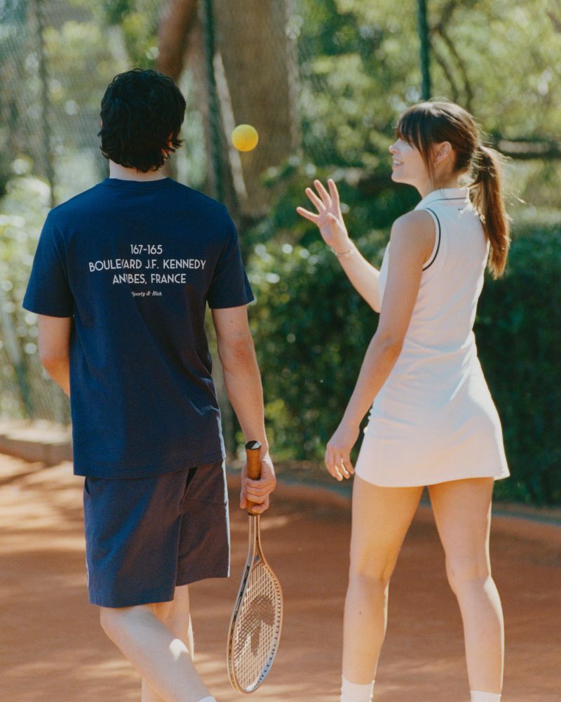 A man and woman in tennis attire stand on a clay court, the woman reaching for a yellow ball, with trees visible in the background.
