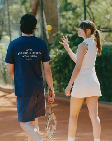A man and woman in tennis attire stand on a clay court, the woman reaching for a yellow ball, with trees visible in the background.