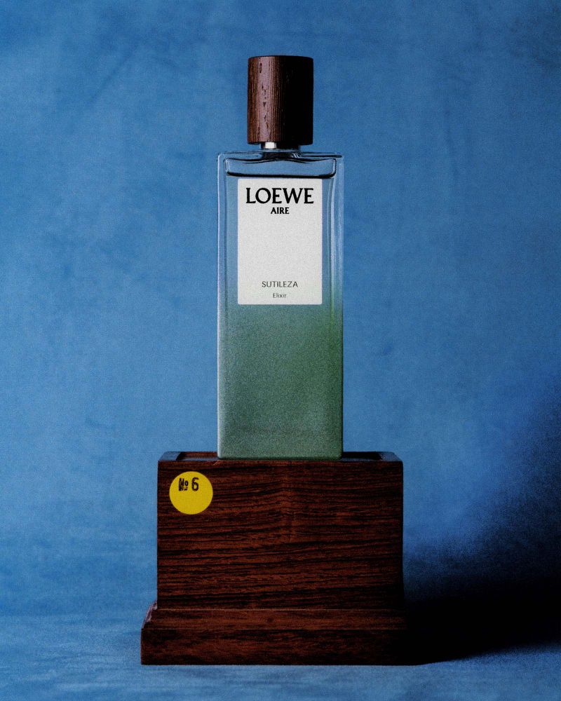 Loewe Aire Sutileza perfume bottle standing on a wooden pedestal against a textured blue backdrop, featuring a yellow circular sticker.