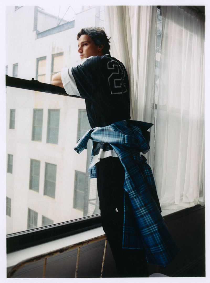 Young man in a black jersey with number 23 standing by a bright window, wearing a blue plaid shirt tied around his waist.